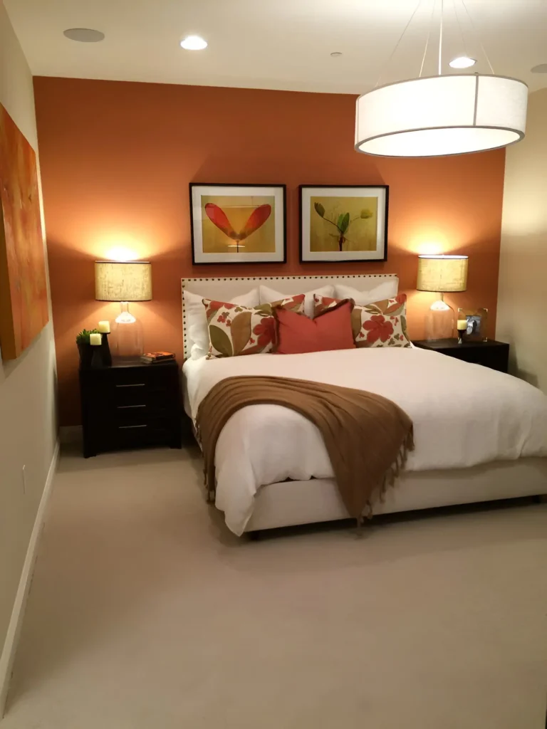 Bedroom with a muted orange accent wall, white bedding, a light brown throw blanket, dark wood nightstands, two framed artworks, and a large drum pendant light.