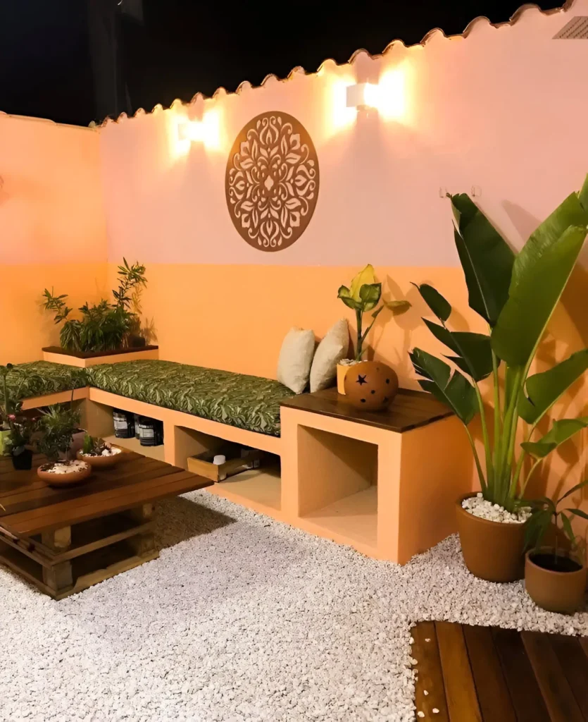 Outdoor lounge area with a two-toned orange wall, a built-in bench with green cushions, white gravel flooring, various potted plants, and a wooden coffee table.