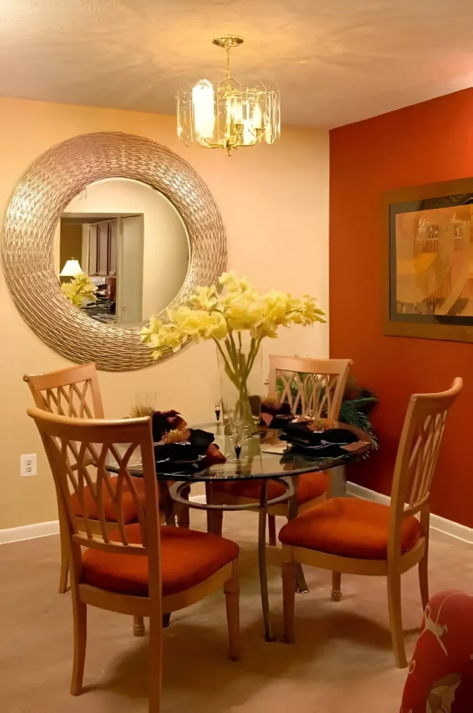 Dining room with a warm orange accent wall, a neutral-colored wall, a round glass table, four chairs with orange cushions, a large round mirror, and a chandelier.