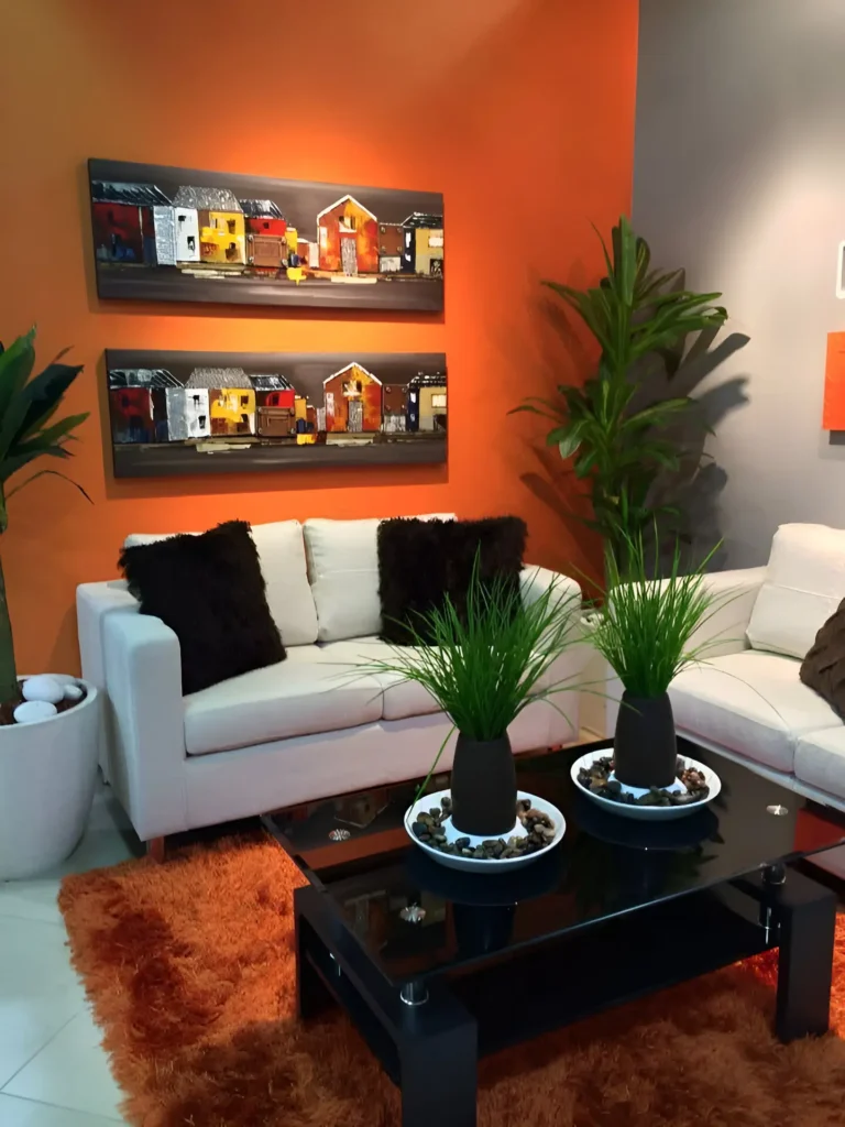 Living room with a warm orange accent wall, a white sofa with dark throw pillows, a black coffee table with green plants, a plush orange rug, and two pieces of artwork featuring buildings with orange accents.