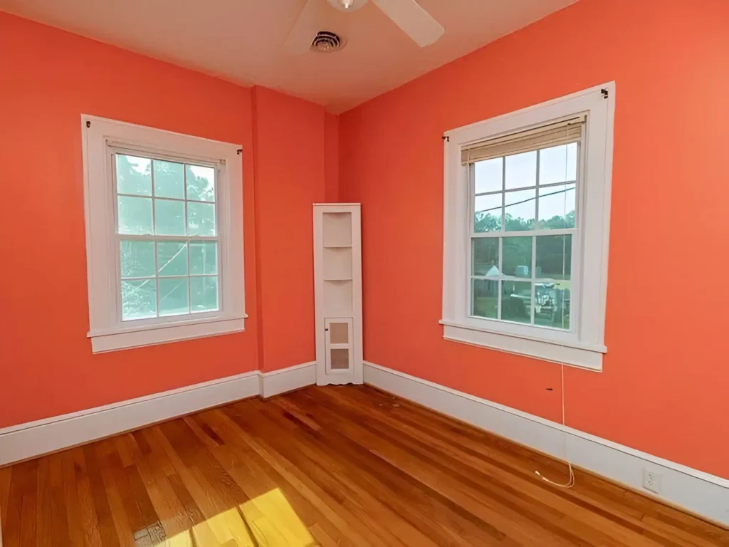 Room with warm orange walls, white trim, two windows with white frames, hardwood floors, and a small built-in white cabinet.