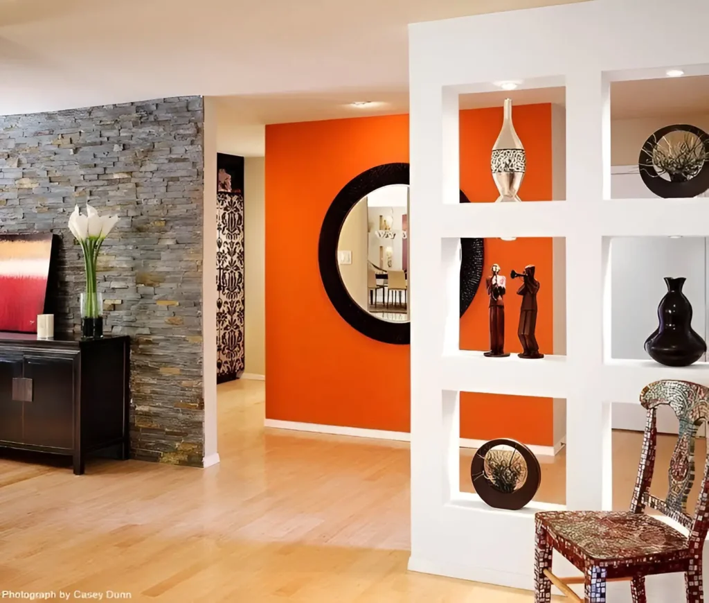 Open-concept living area with a warm orange accent wall, a white partial wall with built-in shelving displaying decorative objects, a light wood floor, and a gray stone accent wall in the background.