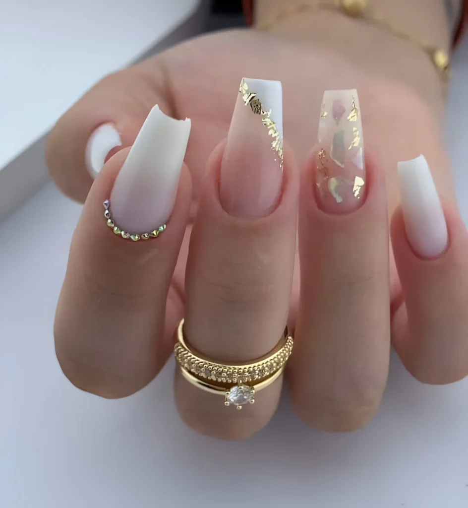 Milky white ombre nails with gold foil and rhinestone accents.