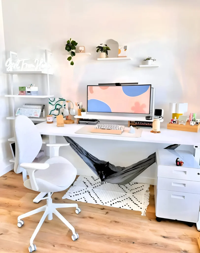 White desk setup with a large monitor, floating shelves, a small hammock, and a patterned rug.