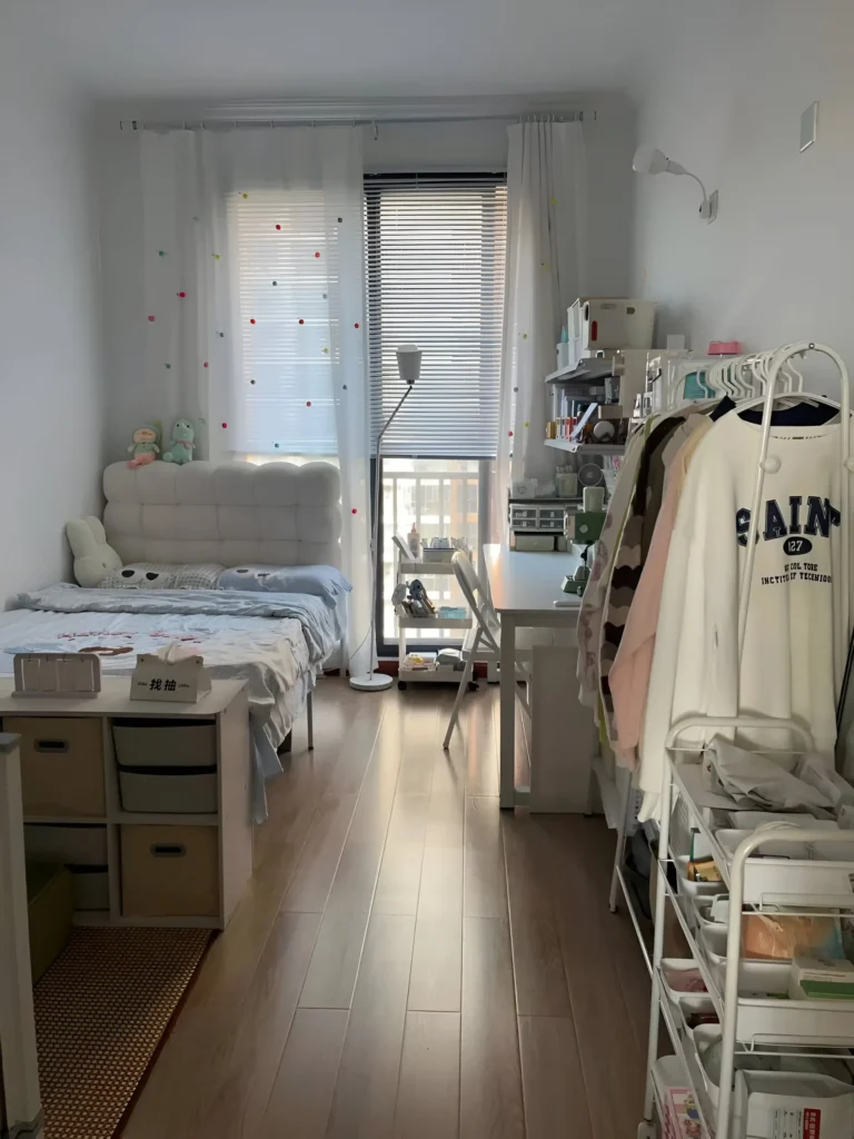 Small bedroom with a tall, white storage unit and clothing rack, utilizing vertical space for organization. The floor is light wood.