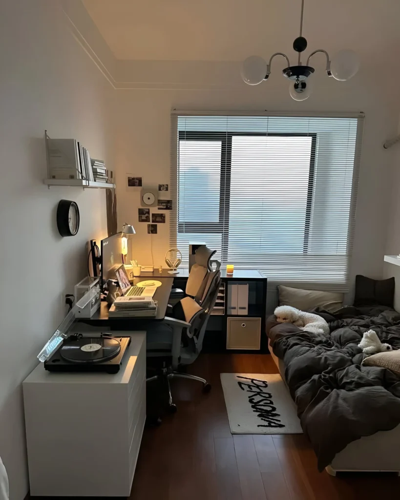 Small bedroom with a desk positioned next to the bed, serving as both a workspace and a nightstand. The room features light walls and dark bedding.