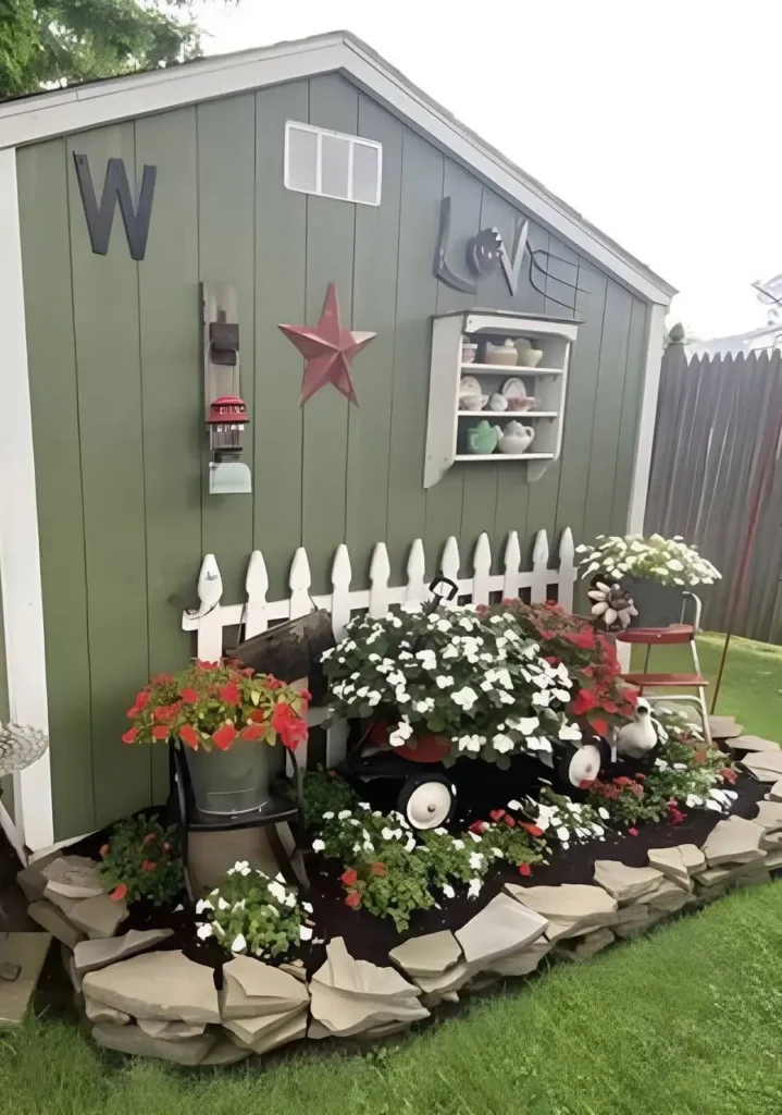 A garden shed decorated with flowers, including impatiens planted in a wagon and containers, surrounded by a small stone border.