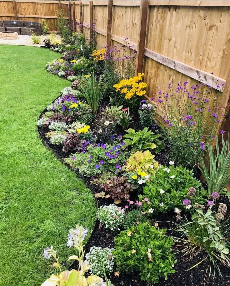 A curving flower bed along a wooden fence, filled with a variety of plants including boxwood, alyssum, and vervain, with dark mulch contrasting against the green lawn. Beautiful Flower Garden Ideas